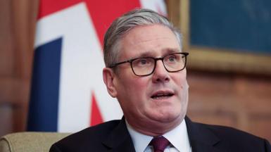 Sir Keir Starmer in a black suit and red tie. He is wearing glasses. The Union Jack is in the background.