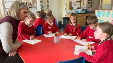 A teacher sits at a red table with a group of young children in a classroom. The children, wearing red school jumpers, draw on paper with coloured pencils. Classroom displays, storage units and children’s chairs are visible in the background.