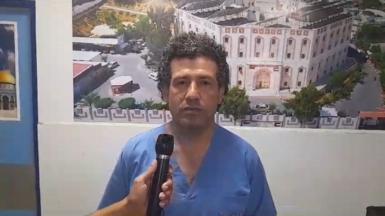Dr Adnan Al-Bursh wearing surgical scrubs speaking into a microphone being held in front of him