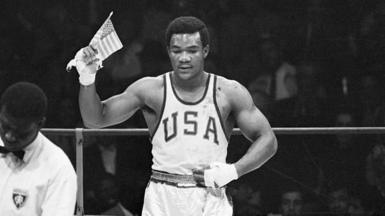 A black-and-white photo of George Foreman in a boxing ring wearing a sleeveless top with USA on it, and holding a USA flag