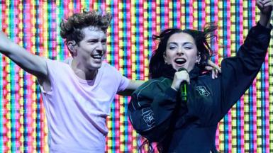 Charli XCX and Troye Sivan on stage, with a multi coloured LED lit background. Troye is on the left, wearing a white short sleeved top and has his left arm around Charli with his right arm outstretched. Charli is on the right wearing a black jacket, singing into a black microphone with her left arm outstretched.