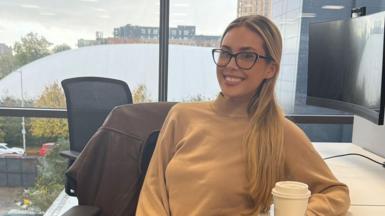 A girl smiling in a office setting at her desk holding a coffee cup wearing glasses