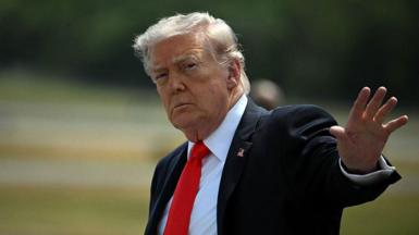 Donald Trump, wearing a suit, raises a hand towards the camera.
