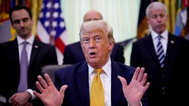 US President Donald Trump wearing a navy suit, white shirt, and gold tie holds both of his hands up as he speaks during an event in the Oval Office of the White House. Two mean in suits and ties are on located on the left and right of him.
