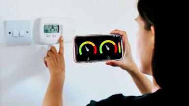 A woman with black hair and a black top looks at a smart meter on her smartphone whilst adjusting the thermostat in her home