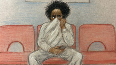 A court sketch of Axel Rudakubana sitting on a red chair. He has black, curly hair and is wearing a grey sweatshirt and joggers. He has his sweatshirt pulled up over his face, only his eyes are visible. 