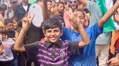 Palestinian children celebrate in Al-Mawasi, Rafah, Gaza Strip, on October 9, 2025, following news of a new Gaza ceasefire deal. A boy at the front of the shot is wearing a black and purple striped top. He is smiling at the camera and making the 'peace' sign with his fingers. 