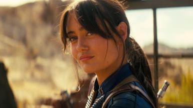 A shot from Fallout shows Ella Purnell in her blue vault suit, hair tied back, looking quizzically at something in the distance. She's surrounded by desert and bathed in a dusky yellow sunlight. 