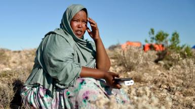 A woman sitting on a mountainside wearing a light grey scarf covering her head and shoulders. She is looking into the distance and holding a mobile phone on her lap.