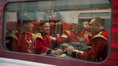 A group of guards sitting on a train with red seats. They are wearing the red uniform with yellow shoulders. They are smiling and chatting, one man has his had on the shoulder of another.