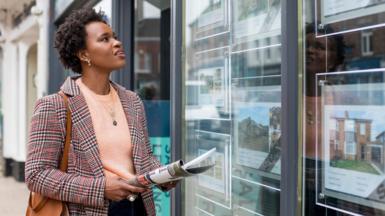 A stock image of a lady perusing an estate agent's available properties in the window. She carries a newspaper, a handbag, and a blazer.