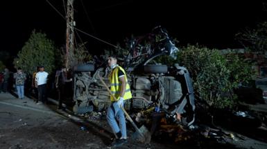 A young man in hi-vis holds a shovel in front of a car, on its side and destroyed by an apparent explosion. Other young men look on. It's dark and the car is on a street in front of a hedge.