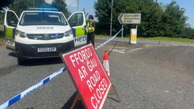A picture of a blue and white police cordon with a red road closed sign and a traffic cone in front. Behind the cordon is a police van with a female officer stood next to it and a traffic sign pointing to Carmarthen.