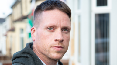 A man with short brown hair and light stubble looks at the camera. Behind him are a row of terraced houses and a Welsh flag. 