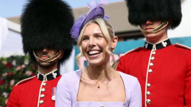 A woman in a lilac dress and fascinator smiles in front of two guardsmen wearing full parade uniform of black busby hats and red jackets with black collars