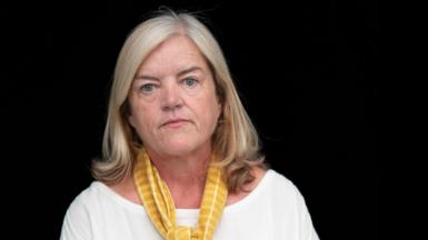 Baroness Louise Casey pictured against a black background, she has shoulder-length blonde hair and wears a white boat-necked top and yellow scarf 