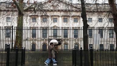 A woman holding an umbrella past black railings in front of the Royal Mint Court, the proposed site of the new Chinese embassy in London.
