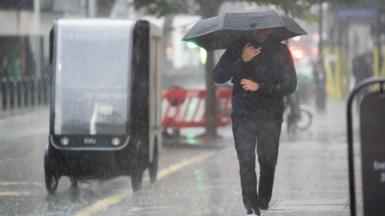 A man wearing a sodden jacket and trousers carries an umbrella down a street through a downpour 