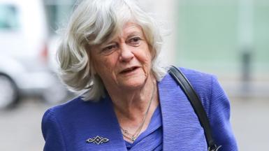 Anne Widdecombe walking wearing a blue suit and with a black leather handbag strap over her shoulder