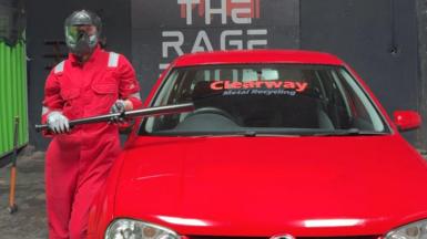 Picture of a woman in a helmet and protective gear that's red. She is holding a baseball bat next to a car.