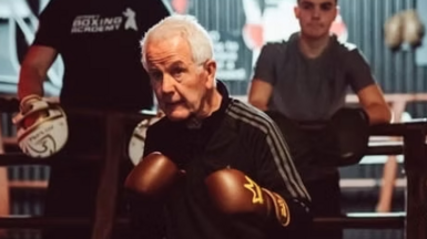 Frank Gilfeather is just outside the ring in a boxing stance wearing a black tracksuit with a white trim. He is also wearing brown boxing gloves with golden crowns on them and behind him are four onlooking students who are standing inside the ring leaning on the ropes.