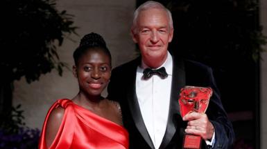 Jon Snow holding a Bafta and  standing with his wife Precious Lunga who wears a red red  