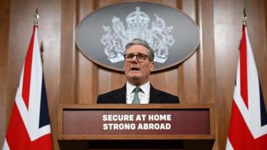 Sir Keir Starmer speaking at a podium flanked by two Union Flags, behind a podium bearing a sign which reads "Secure at home, strong abroad"