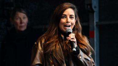 A woman in a brown leather jacket and brown hair holds a microphone