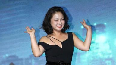 South Korean comedian Park Na-Rae with a short black bob and a black dress waving against a blue background at a press event