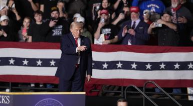 US President Donald J. Trump stands in front of stars and stripes bunting with a crowd clapping and taking photos w phones behind him inside an auditorium. He is in a suit and pointing