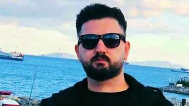 Kardo Ranya stands in the foreground, wearing dark sunglasses, a black jacket, and a dark shirt. Behind him is the sea, a few ships are visible near the horizon. 