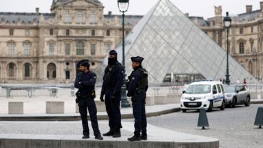 Three police officers stand on concrete with the Louvre glass pyramid in the background