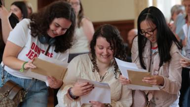 Photo shows three girls collecting their GCSE results. They all have long, dark hair and are smiling down at their results papers. One of the girls wears a t-shirt which says 'don't be jealous', one is a wheelchair user and one is wearing glasses.
 
 