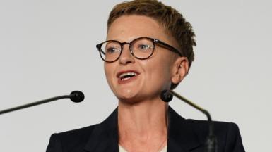Bev Craig has short spiky red hair, dark glasses and wears a dark jacket with white roll-neck top.