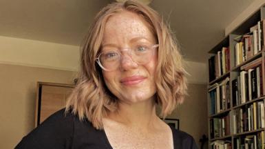 A smiling Laura Etchells wearing glasses and smiling, standing in her living room with bookshelves seen on the right