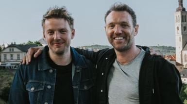 Two men stand, smiling, with a view of a traditional European city spreading out behind them. The man on the right, wearing a black hoodie over a grey t-shirt, drapes his right arm over the shoulder of the other man, who's wearing a blue denim jacket over a black t-shirt.