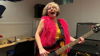 Janet is in a music studio playing her guitar and giving a big laugh with her head back, wearing a bright pink fluffy gilet 