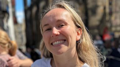 Courtney Buchner. Courtney is a woman in her 20s with long blonde hair worn loose. She has a nose ring and smiles at the camera. She's pictured outside in Edinburgh on a busy street, wearing a white T-shirt. 