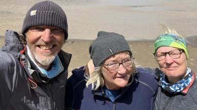 Craig, Sophie and Rona are in the outdoors wearing and smiling for a picture Craig is taking on his phone. Craig and Sophie are wearing fleece hillwalking hats while Rona has colourful scarf around her head.