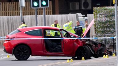 A red car that has crashed has police tape around it. Police officers in the background are wearing neon yellow hi-viz jackets 