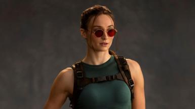 A photo shows Sophie Turner as Lara Croft. She wears red-tinted sunglasses, her hair tied back. She wears a green vest top and tight, thigh-length shorts. One of her fingerless-gloved hands rests on a pistol in a thigh holster.