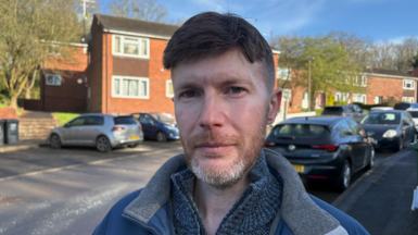A man stood in a street with cars behind him and a row of houses. He has brown hair and greying beard. He's wearing a blue jumper and jacket