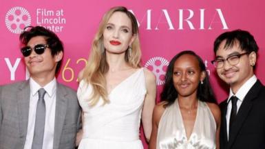 An image of Angelina Jolie wearing a white dress and three of her children on the red carpet standing in front of a pink board 