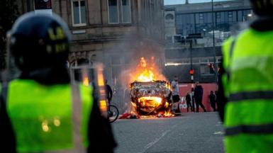 Police officers wearing riot gear in Sunderland, viewed from behind close to the camera, as in the middle distance an overturned car burns and a crowd mills around