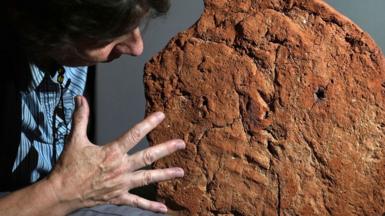Helen Strudwick on the left with the base of an ancient Egyptian soul house. She is side on and only the top of her dark hair and her nose can be seen as she looks a the soul house on the right. She has stretched out her right hand towards the artefact. The base of the house is in lumpy light brown clay. On its base, towards its bottom side, can be seen light ridges of fingers. 
