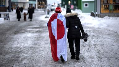 Two people walk down an icy street. One is wearing a draped Greenland flag.
