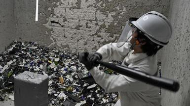 Picture of two people in a rage room smashing items with baseball bats. They are wearing hard hats, visors and white overalls.