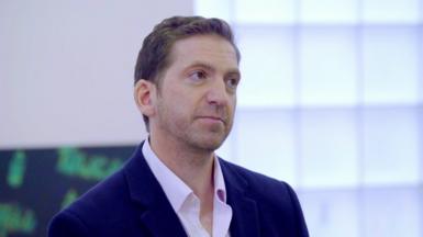 Dr Faisal Maassarani has short brown-blonde hair, is wearing a blue suit and an open-necked pink shirt. He is standing in his surgery in a clip from a BBC health programme called The Truth About Carbs in 2018. 