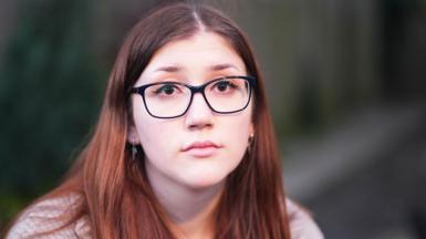 A close-up portrait of Laura Kenny, facing the camera directly. She has long brown hair that falls past her shoulders and she is wearing dark-framed glasses. Her expression is calm and neutral.