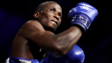 Nuhu Batte, with blue boxing gloves on and with an arm flexed, preparing to fight.